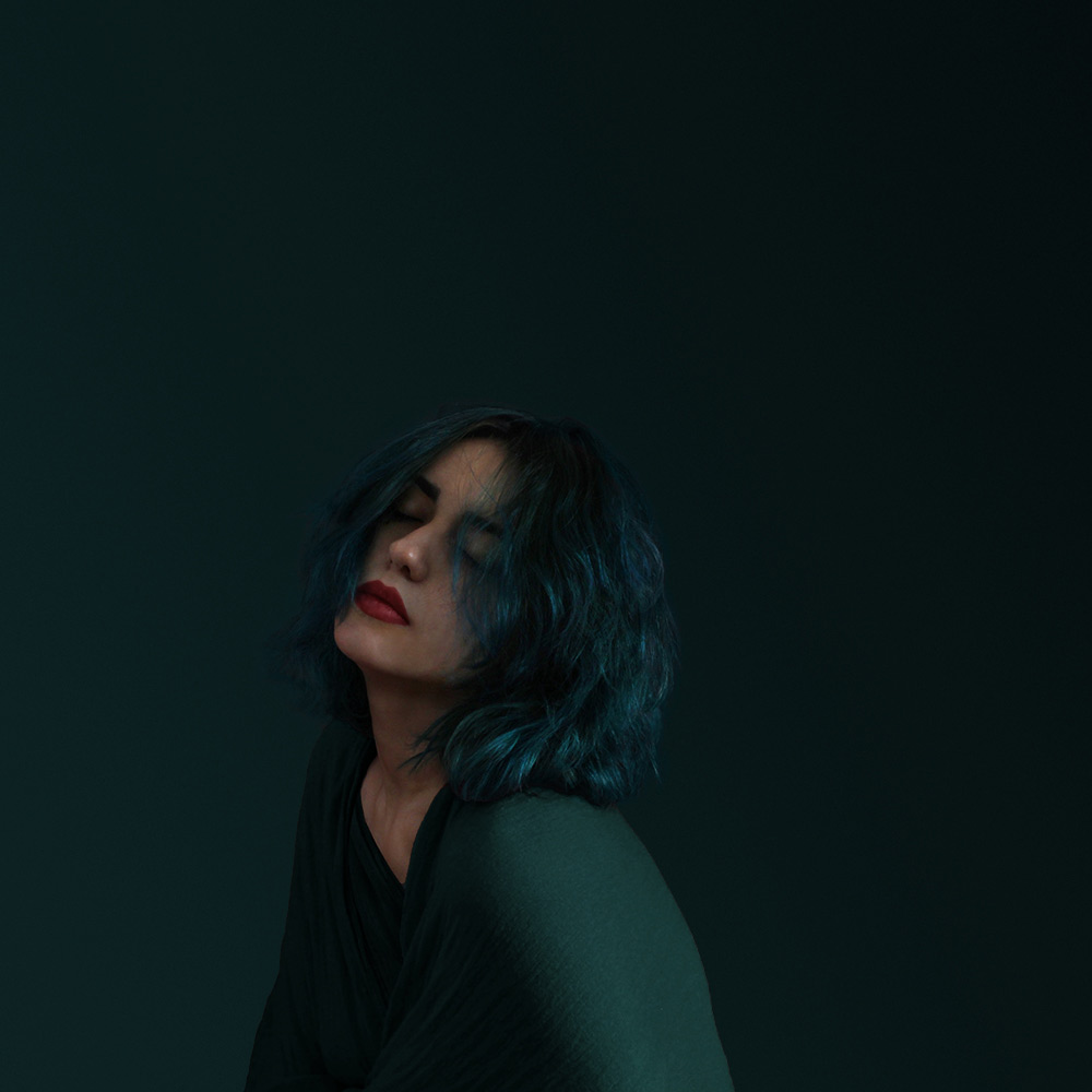 Portrait of a woman with blue hair and eyes closed against a dark teal background