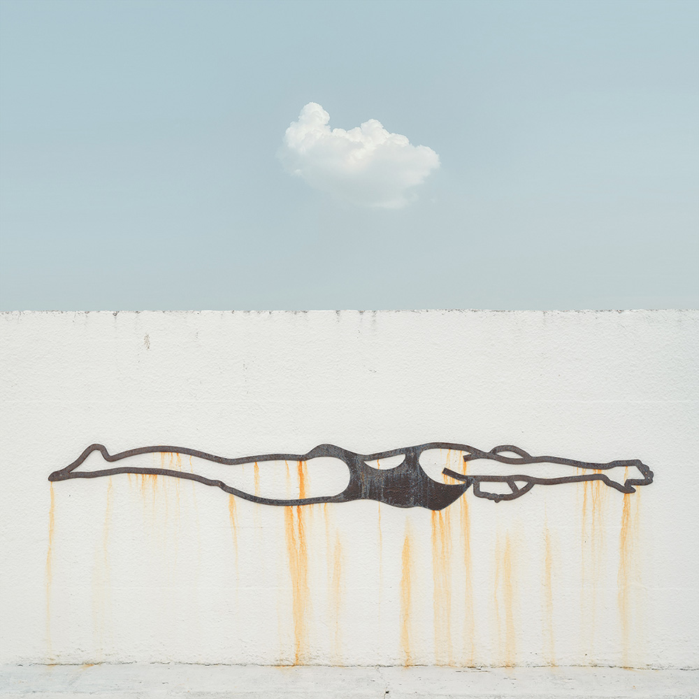 A rusted metal swimmer sculpture on a white wall under a single cloud